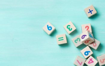 A scattered collection of wooden toy blocks featuring colourful numbers and mathematical symbols, set against a light turquoise wooden background.