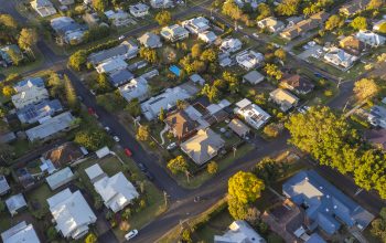 An aerial bird's-eye view of a suburban neighborhood at golden hour, showing residential houses with varied rooflines, swimming pools, and cars parked along tree-lined streets.