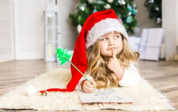A young girl wearing a Santa hat lies on a fuzzy rug, thoughtfully looking away while holding a green pencil over a colouring page.