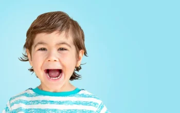 A young boy with brown hair in a striped blue and white shirt smiles widely with his mouth open against a solid light blue background.