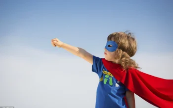 A young child wearing a blue mask and a red cape extends their arm with a clenched fist, posing as a superhero against a clear blue sky.